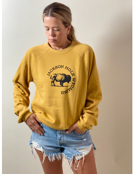 A person wearing a mustard sweatshirt with 'Jackson Hole' and a bison graphic on it, standing against a neutral background.