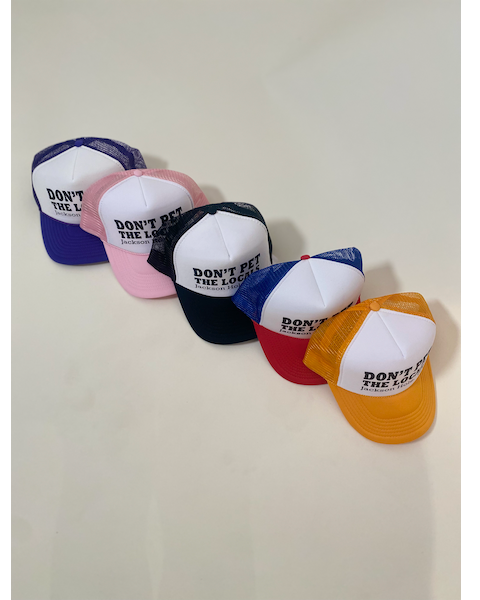 A series of baseball caps displayed in a row with the text 'DON'T PET THE LOCALS' on them. The caps are in a line with colors red, pink, blue, white, yellow, and green.