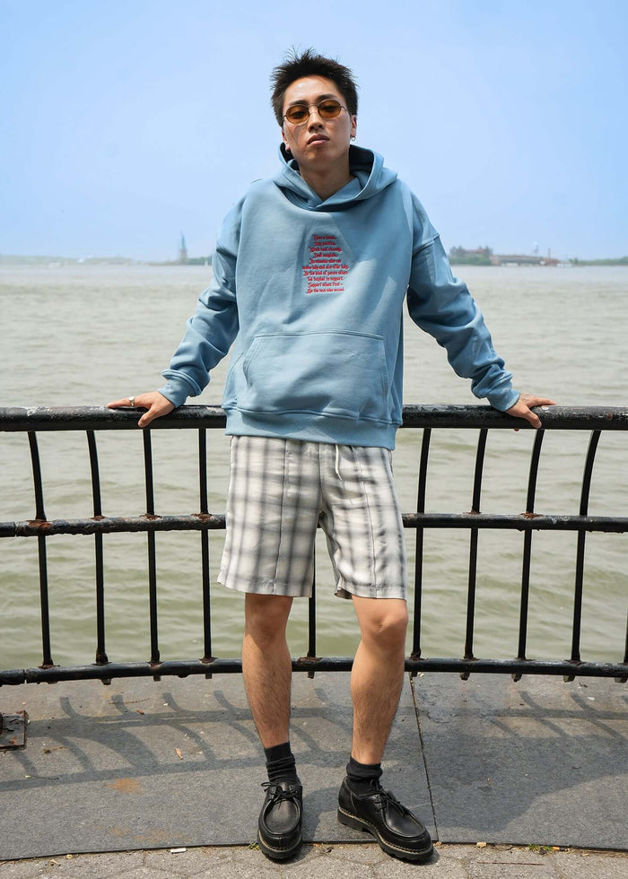 Model wearing Words Of Wisdom lightweight terry hoodie by Éclectique near water, featured with stylish plaid shorts.