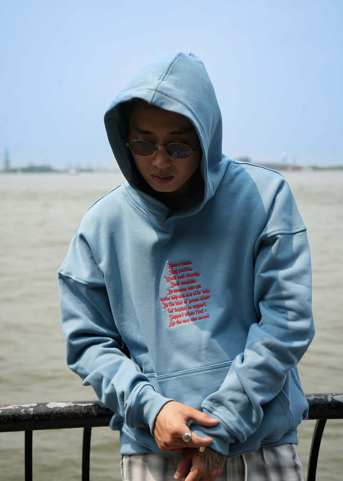 Man wearing a lightweight terry hoodie with embroidered words of wisdom, standing by a waterfront.