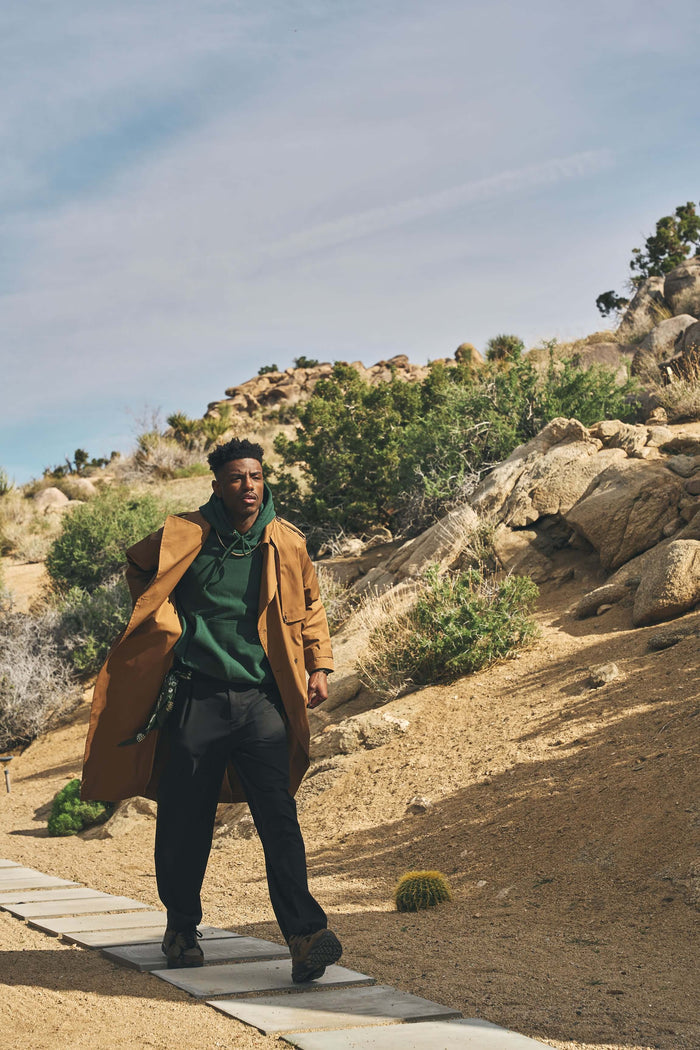 Man wearing a stylish Men’s Camel Trench Coat while walking in a desert landscape with rocky terrain.