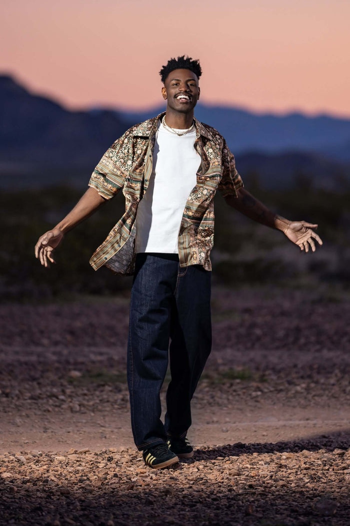 Man wearing men's premium denim jeans with a patterned shirt in a desert landscape during sunset.