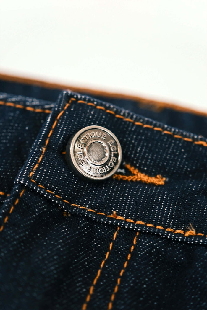 Close-up of a metal button on men's premium denim jeans with dark wash fabric and contrasting stitching.