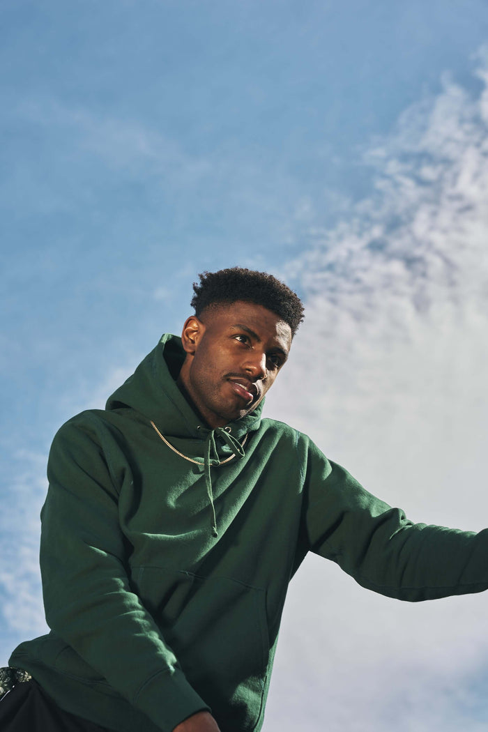 Young man wearing a mid weight cotton hoodie in ivy green under a blue sky.