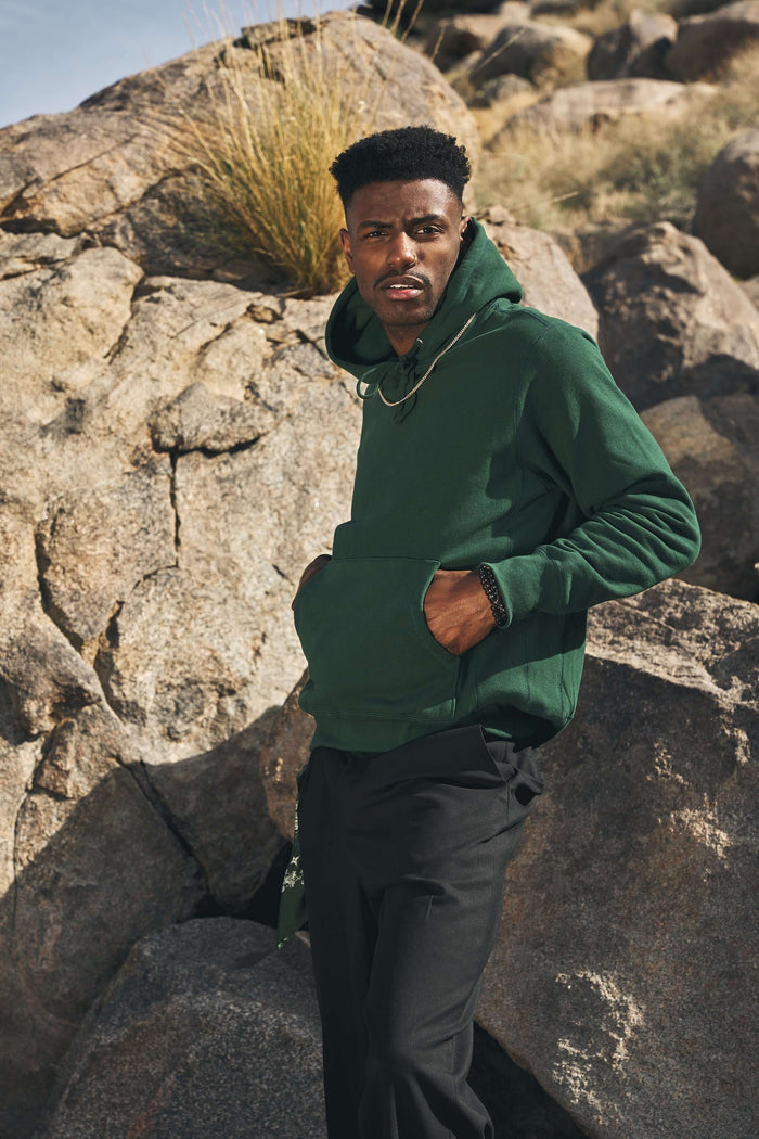 Model wearing a mid weight cotton hoodie in ivy green, featuring a pouch pocket and posed against rocky terrain.