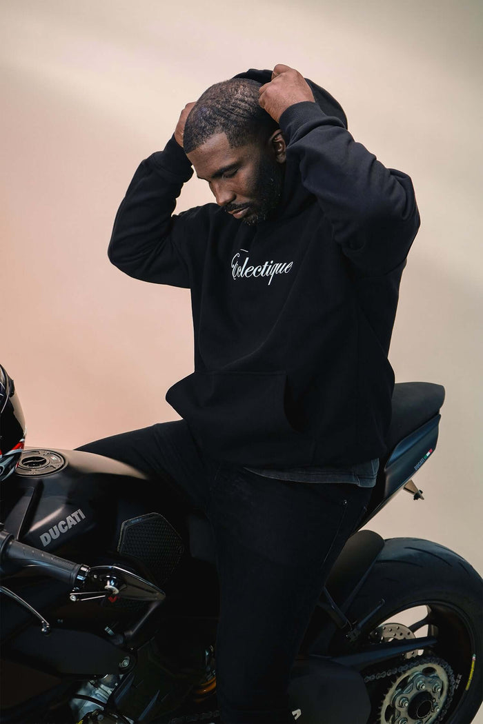 Man wearing a black midweight cotton hoodie with Éclectique script logo while sitting on a motorcycle.