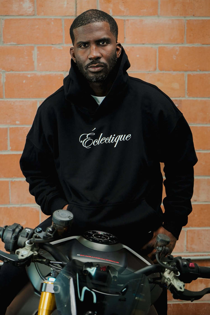Midweight cotton hoodie in black with Éclectique script logo, model standing with motorcycle, stylish and modern look.