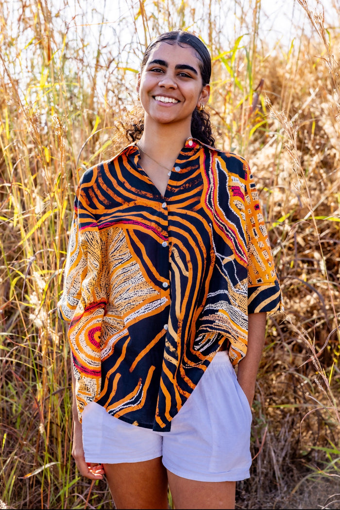 A woman smiling outdoors in front of dry grass, wearing a short-sleeve button-up shirt with a Aboriginal artwork pattern in orange, black, white, and red, paired with white shorts.