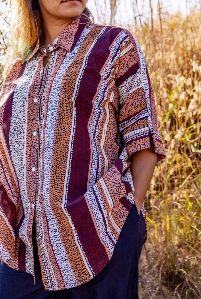 A woman wearing a short-sleeve button-up shirt with vertical stripes Aboriginal artwork in burgundy, brown, orange, and white, paired with dark navy skirt, set against a background of dry grass.